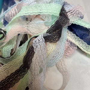May include: Close-up of a pile of colorful lace ribbons in various pastel shades. The ribbons include light blue, green, pink, white, and black, with a delicate, scalloped edge. Ideal for crafting and sewing projects.