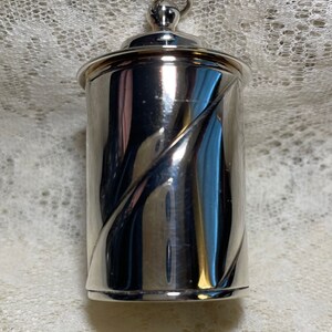 May include: A cylindrical silver-toned metal container with a polished, reflective surface. The body features a distinct diagonal recessed groove wrapping around the exterior. It has a flat base and a rounded lid topped with a small metal loop.