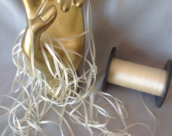 May include: A spool of off-white satin ribbon is wound around a black plastic spool. The ribbon is also wrapped around a gold-colored hand sculpture.