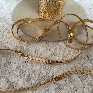 Puede incluir: Cinta de adorno de lentejuelas doradas. La cinta está hecha de pequeñas lentejuelas redondas cosidas juntas sobre un soporte delgado y flexible. Las lentejuelas son de color dorado brillante y la cinta tiene aproximadamente 1,2 cm de ancho.