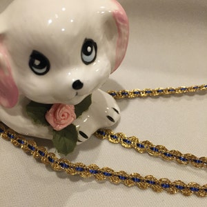 May include: A white porcelain figurine of a dog with pink ears and a rose on its neck. The dog is wearing a gold and blue trim ribbon around its neck.