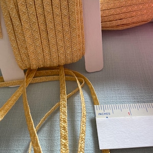 May include: Close-up of a spool of yellow raffia ribbon with a woven texture. The ribbon is approximately 2.5 cm wide. A ruler is placed next to the ribbon for measurement.