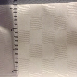 May include: A close-up of a white fabric with a checkered pattern. The fabric is textured with diagonal lines. A ruler is visible on the left side of the image, measuring up to 3 inches.