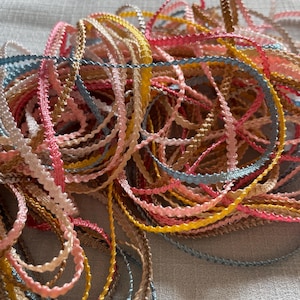 Op de afbeelding: Een stapel kleurrijke decoratieve sierbanden in verschillende tinten roze, blauw, geel en beige. De sierband heeft een geschulpte rand en is strak opgerold, waardoor een textuur ontstaat. Geschikt voor knutsel- en naaiprojecten.