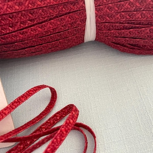 Puede incluir: Primer plano de un rollo de ribete trenzado rojo, sujeto con una banda de plástico blanco. Un trozo del mismo ribete rojo se encuentra en primer plano, mostrando su textura tejida. Ideal para proyectos de costura o manualidades.