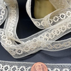 May include: A close-up of a delicate, off-white lace trim with a repeating floral pattern. The lace is shown next to a US penny for scale.