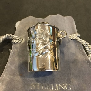 Puede incluir: Un recipiente cilíndrico de plata con un diseño floral ornamentado. El recipiente tiene un lazo en la parte superior para colgarlo. El recipiente está sentado sobre una superficie de terciopelo gris con las palabras "Sterling Silver" impresas en él.