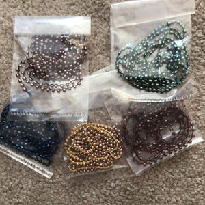 May include: Five bags of ball chains in various colors: dark blue, bronze, gold, dark purple, and green. Each chain is in a clear plastic bag. The chains are on a neutral-colored carpet.