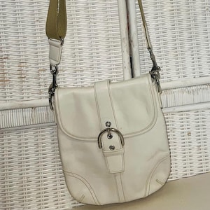 May include: Off-white leather crossbody bag with a flap closure and silver-tone hardware. The bag has a long olive green strap and a buckle detail on the front. The bag is hanging against a white wicker background.
