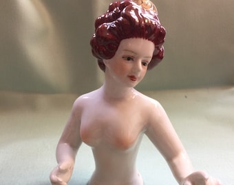 May include: A porcelain doll head with a white body and red hair. The doll has a hole in the bottom of the torso, suggesting it is a vintage doll head for crafting.