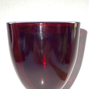 Könnte beinhalten: Ein rubinrotes Weinglas mit rundem Kelch und kurzem, spiralförmigem Stiel. Das Glas hat einen kreisförmigen Fuß und besteht aus einem glänzenden, transparenten Material. Das Glas ist leer und steht auf einer weißen Oberfläche.