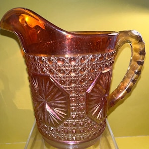 May include: A vintage iridescent glass pitcher with a diamond pattern and a starburst design. The pitcher has a handle and is a light amber color.