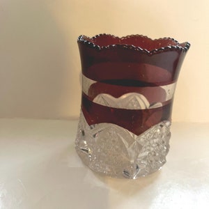 May include: A ruby red glass vase with a scalloped rim and a clear, cut-glass base. The vase features a decorative band of clear glass and a detailed, geometric pattern on the base. The vase is approximately 4 inches tall.