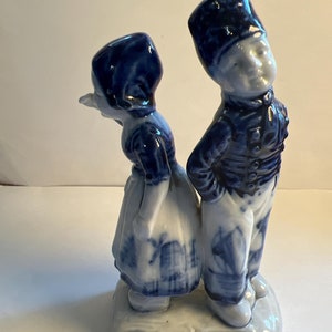 Puede incluir: Una figurita de porcelana azul y blanca de una pareja holandesa. La mujer lleva un vestido azul y blanco y un gorro azul. El hombre lleva un uniforme azul y blanco y un sombrero azul. La figurita está de pie sobre una base blanca.