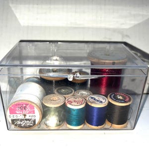 May include: A clear plastic box containing various spools of thread in colors like red, blue, black, and green. Some spools have labels with text. The box also holds several small, round, wooden spools and a clear, domed object.