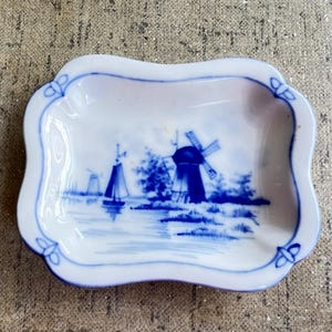 May include: A rectangular porcelain trinket dish with scalloped edges, featuring a blue and white hand-painted scene of a traditional windmill and a sailboat on water. The border is decorated with blue floral-like motifs at each corner.