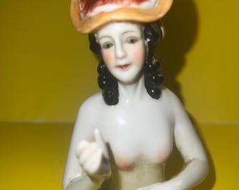 May include: A porcelain figurine of a woman with a brown and yellow hat. The woman is wearing a white dress and has black hair.