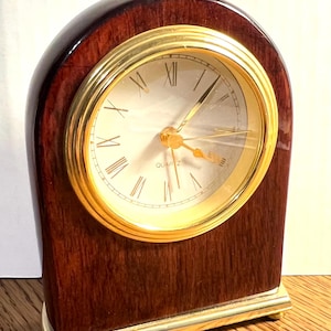 Peut inclure: Une horloge à quartz en bois avec une lunette dorée et des chiffres romains. L'horloge a un boîtier en bois foncé et arqué et une base dorée. Le cadran est blanc cassé.