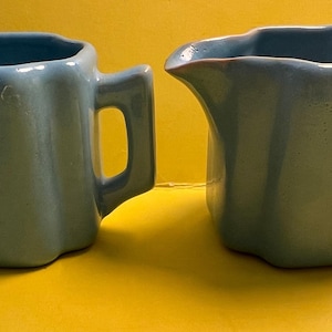 May include: A set of two blue ceramic creamer and sugar containers with a geometric design. The containers have a fluted design and a handle.