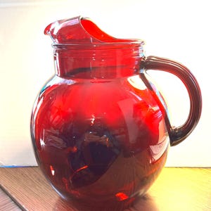 Puede incluir: Una jarra de vidrio rojo rubí con cuerpo redondeado y asa curva. La jarra tiene un pico vertedor para facilitar el servicio. El vidrio es liso y reflectante, con un acabado brillante. Probablemente se utiliza para servir bebidas.