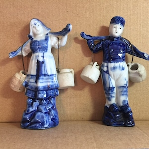 Puede incluir: Dos figuritas de porcelana azul y blanca de un niño y una niña que llevan cubos en un palo sobre sus hombros. Las figuritas son de estilo holandés tradicional.