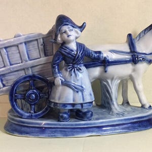Puede incluir: Una figurita de cerámica azul y blanca de una niña con un vestido tradicional holandés tirando de un carro con un burro. La figurita está pintada a mano y tiene un diseño detallado.