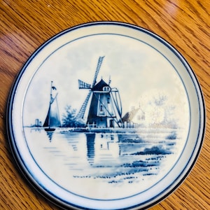 Puede incluir: Plato redondo de cerámica blanca con una escena de molino de viento azul y blanco. El plato presenta una ilustración detallada de un molino holandés, un velero y reflejos de agua. El borde tiene un borde azul.