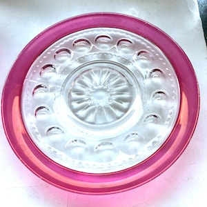 May include: A clear glass plate with a vibrant pink rim. The plate features a central design and a ring of decorative indentations. The pink rim adds a pop of color to the otherwise transparent plate.