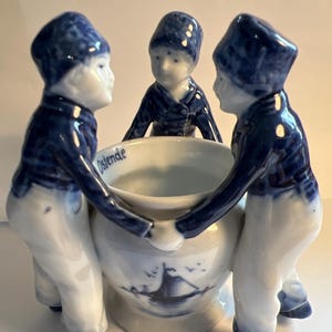 Puede incluir: Figuras de cerámica azul y blanca de cuatro niños que llevan una olla grande con una escena de molino de viento. La olla está inscrita con la palabra "Ostende".