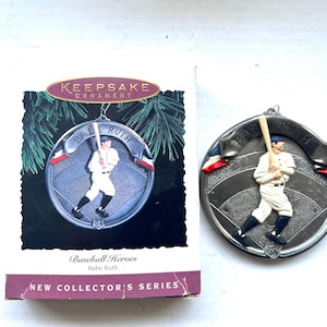 May include: A silver-colored Babe Ruth baseball ornament with a baseball player figure in a white uniform, holding a bat. The ornament is round with the words "Babe Ruth" and "94" on it. The ornament is part of the "New Collector's Series".