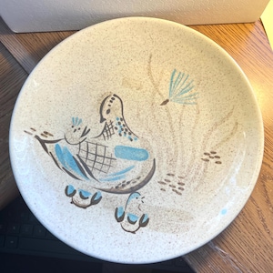 Puede incluir: Un plato redondo de cerámica moteada con un diseño moderno de mediados de siglo. El plato presenta un motivo estilizado de pájaro y polluelo en tonos azules y marrones. El plato tiene un diámetro de aproximadamente 25 cm.