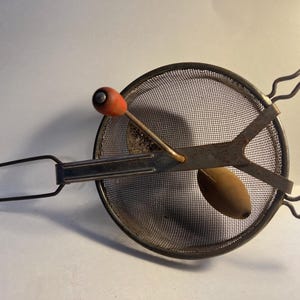 May include: Vintage food mill with a metal mesh strainer, a wooden oval-shaped piece, and a red knob. The handle and frame are made of metal. This kitchen tool is designed for straining and pureeing food.