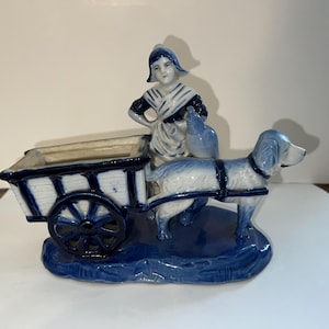 Puede incluir: Figura de cerámica azul y blanca que representa a una mujer, un perro y un carro. La mujer viste un atuendo tradicional y está junto al carro. El perro está enganchado al carro. La figura está sobre una base azul.
