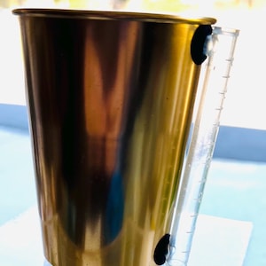 May include: A gold-colored metal measuring cup with a clear plastic handle attached with black fasteners. The handle has measurement markings. The cup is sitting on a white surface.