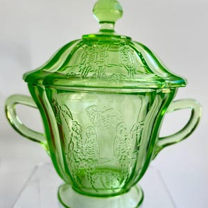 Könnte beinhalten: Eine grüne Zuckerdose aus Glas mit Deckel und zwei Griffen. Die Schüssel und der Deckel weisen ein geprägtes Design von Vögeln und Blättern auf. Das Glas hat ein helles, durchscheinendes Aussehen, und die Schüssel steht auf einem kleinen Sockel.