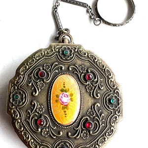 May include: A round, antique-style metal compact mirror with a chain and finger ring. The surface features intricate embossed scrollwork, inset red and green stones, and a central oval enamel inlay depicting a pink rose on a yellow background.