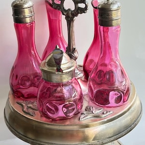 May include: An antique silver-plated serving set with pink glass bottles and a central handle. The set includes four bottles with silver lids and a small jar with a lid, all resting on a decorative silver tray.