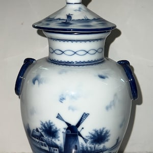Puede incluir: Jarrón de cerámica blanco y azul con tapa, con un diseño de molino de viento y paisaje. El jarrón tiene dos asas y un patrón decorativo alrededor del cuello. La tapa tiene un pequeño diseño de velero.