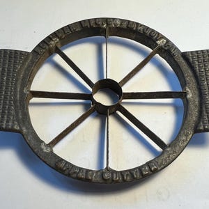 May include: Antique cast iron apple cutter with a circular design. The cutter has a central ring, radiating blades, and two rectangular handles with a grid pattern. The words "APPLE CUTTER" are embossed on the rim.
