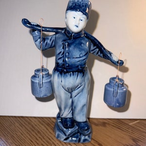 Puede incluir: Una figurita de porcelana azul y blanca de una persona que lleva dos cubos en un yugo. La persona lleva un sombrero azul y un uniforme azul. La figurita está de pie sobre una base.