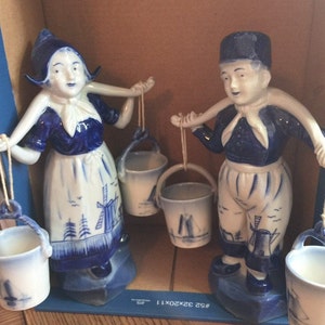 Puede incluir: Dos figuritas de porcelana azul y blanca de una niña y un niño con ropa tradicional holandesa. La niña lleva un cubo y el niño lleva un cubo y un palo largo. Las figuritas están sobre una caja azul y blanca con el texto "# 92 32X20X11"