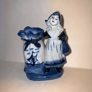 Puede incluir: Figurilla de porcelana azul y blanca de una niña con un vestido tradicional holandés que sostiene una bolsa. La bolsa está decorada con un velero y tiene un cordel.