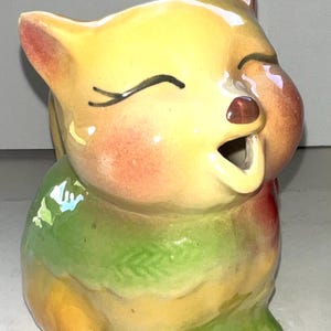 Puede incluir: Figura de cerámica vintage de un gato sonriente con tonos amarillos, verdes y melocotón. El gato tiene los ojos cerrados, una nariz roja y un collar verde. Un artículo decorativo caprichoso.