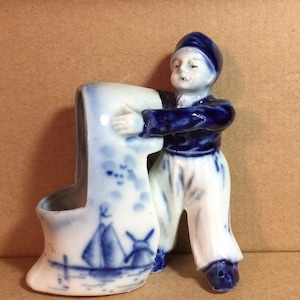 Puede incluir: Una figurita de cerámica azul y blanca de una persona sosteniendo un zapato de cerámica azul y blanca. El zapato tiene un molino de viento y un velero pintados en él.