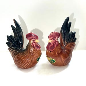 May include: A pair of ceramic rooster salt and pepper shakers. The roosters are brown with black tails and red combs. The shakers have a green "Made in" label on the bottom.