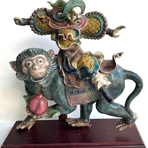 Op de afbeelding: Een gedetailleerde, handgeschilderde keramische sculptuur die een krijger op een aap afbeeldt. De krijger draagt een sierlijk harnas en een helm, terwijl de aap een perzik vasthoudt. De sculptuur is gemonteerd op een donkere houten voet.