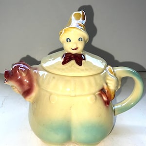 Puede incluir: Una tetera de cerámica caprichosa, amarilla con base azul claro. La tapa presenta una cara sonriente con pajarita roja y sombrero dorado. El pico es una cabeza de cerdo y el asa es verde claro. Un artículo decorativo encantador.