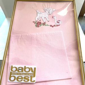 May include: A framed pink baby bedding set with a gold trim. The set includes a pink embroidered fabric with a lamb and duck design, and a matching pink square fabric. The label reads "baby best".