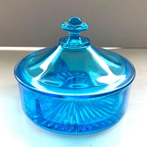 May include: A vibrant, turquoise-colored glass candy dish with a lid. The dish features a round base with a radial pattern and a matching lid with a decorative floral-shaped handle. The glass has a glossy finish.