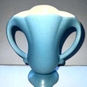 May include: A vintage ceramic vase in a light blue colour with a cream-coloured rim. The vase features two curved handles and a pedestal base. The design is reminiscent of mid-century modern pottery.